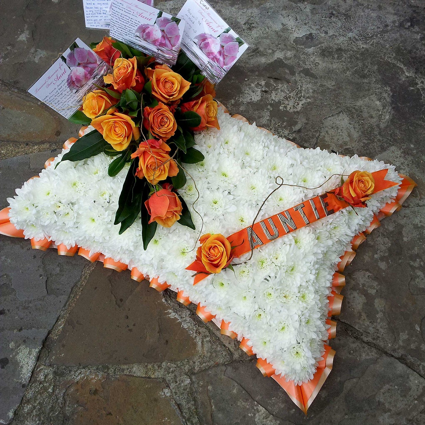 The Cushions & Pillows - White Base by ReCreation Flowers features white seasonal blooms in a cushion shape, accented with orange roses, a ribbon labeled AUNTIE, and condolence cards arranged on a stone surface.