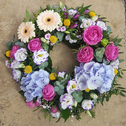 The ReCreation Flowers Funeral Flower Wreath features pink roses, pale gerbera daisies, yellow billy balls, blue hydrangeas, white lisianthus, and green leaves, arranged on a neutral textured background.