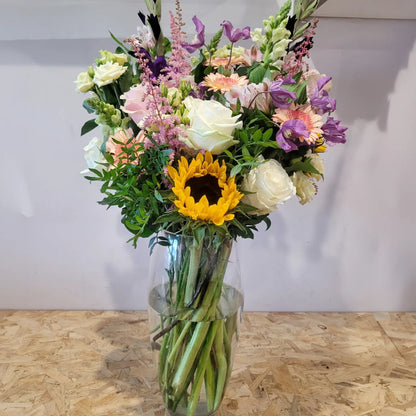 Scent With Love by ReCreation Flowers features a sunflower, white roses, pink and purple blooms, and greenery in a clear glass vase—lovingly arranged by your Leigh-on-Sea florist. Perfect for same-day delivery. Displayed on wood against a light wall.