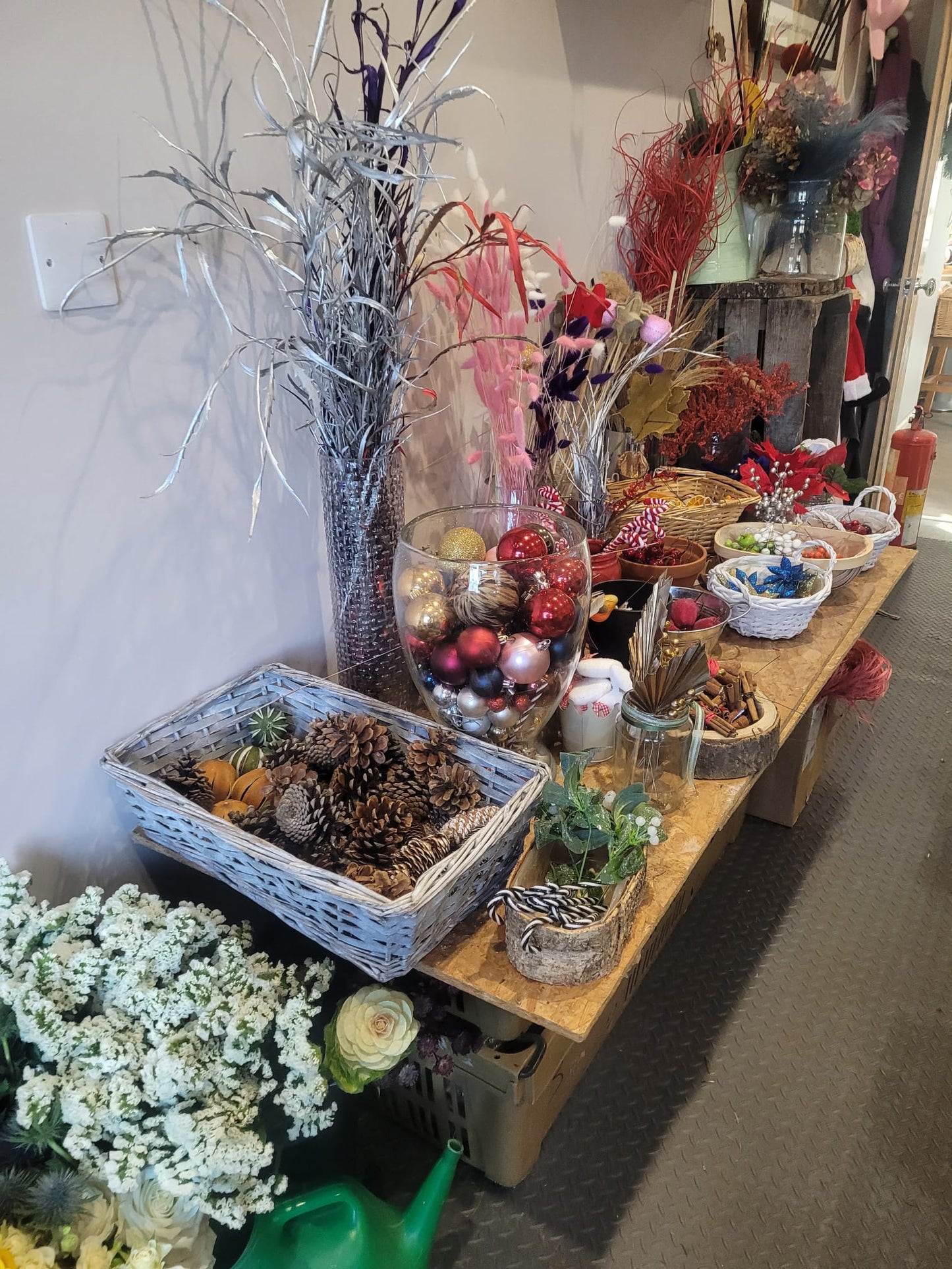 Discover the festive setup for the Christmas Wreath Class by ReCreation Flowers: a table adorned with bowls of pinecones, glass vases filled with ornaments, baskets, dried flowers, and greenery. Nearby, white flowers and a green watering can sit on the floor beside the display.