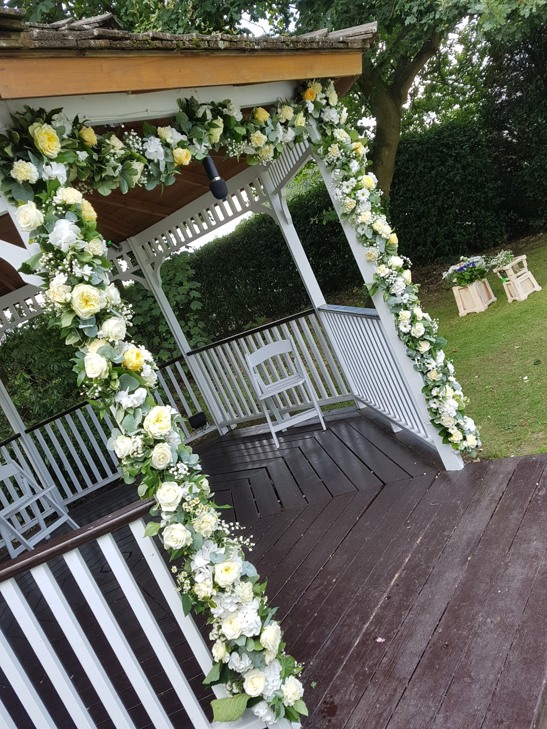 A white gazebo decorated with garlands of white and yellow flowers on its posts stands on a wooden deck in a green garden. Two white chairs sit inside, and flower-filled crates are visible on the grass nearby.