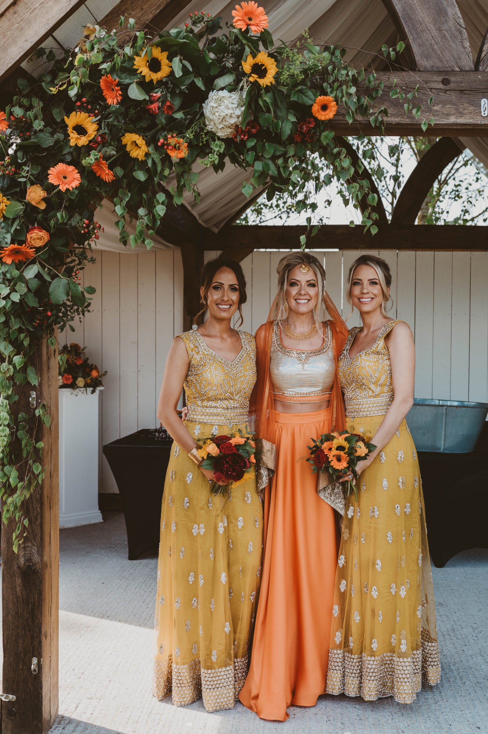 Three women in colorful dresses pose under a wooden arch decorated with orange and yellow flowers. The woman in the center wears an orange outfit; the women on either side wear matching yellow dresses and hold bouquets.