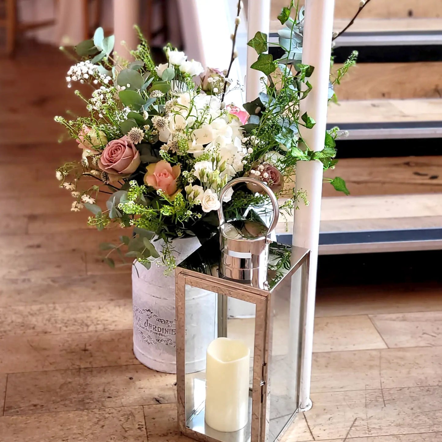 The ReCreation Flowers Beautiful Blooms Package features wedding florals and greenery in a rustic bucket with a metal lantern and candle at the base of a wooden staircase, creating charming venue decor.