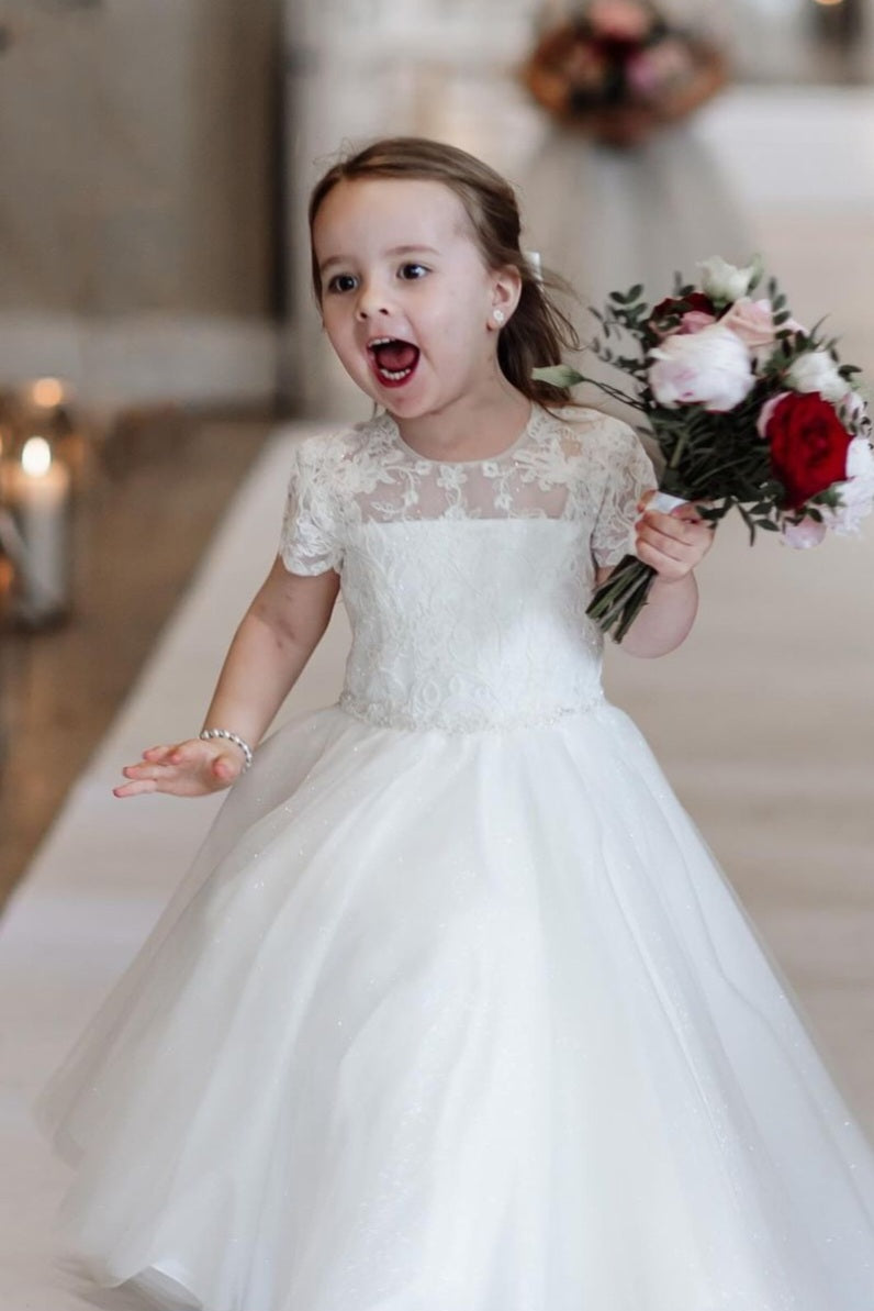 A young girl in a white lace dress excitedly runs while holding a bouquet of flowers. She has a joyful expression, with her mouth open as if shouting or laughing, and is dressed like a flower girl at a wedding.