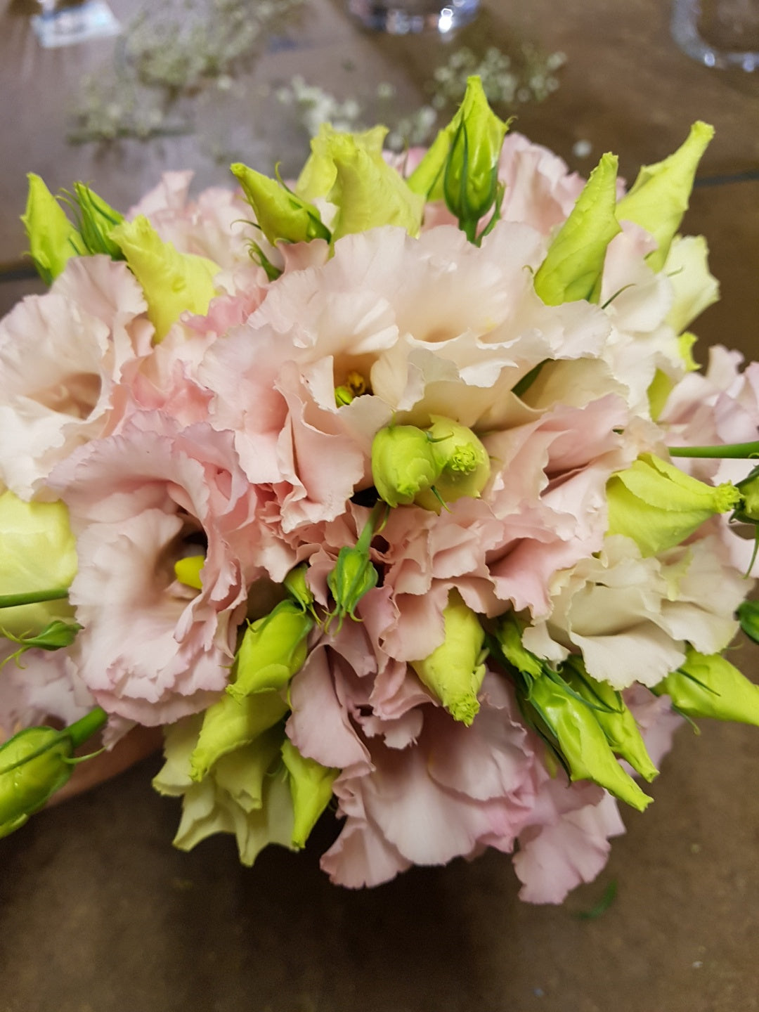 A close-up of a bouquet featuring pale pink lisianthus flowers with light green buds, arranged in a round, compact cluster on a brown surface.
