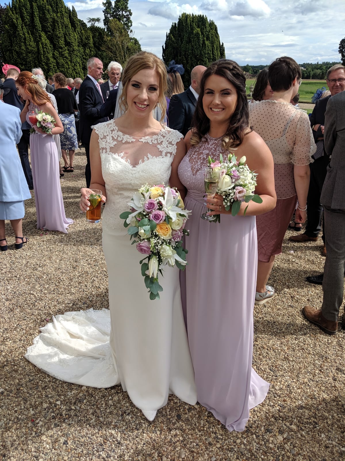 A bride in a white gown and a bridesmaid in a lavender dress stand smiling together outdoors, holding bouquets and drinks, surrounded by guests at a wedding.