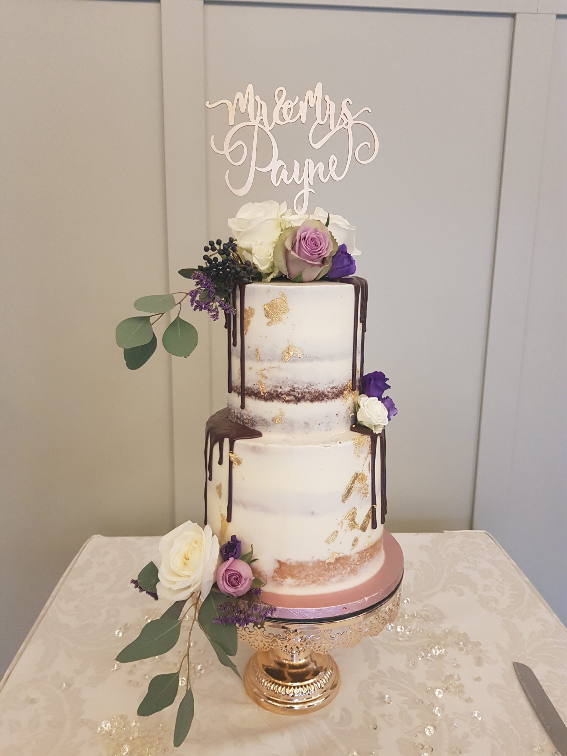 Two-tier semi-naked wedding cake decorated with white and purple flowers, gold leaf accents, chocolate drip, and a Mr & Mrs Payne topper, displayed on a gold stand with a floral tablecloth.