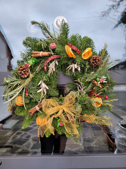 A Christmas Wreath Class by ReCreation Flowers, featuring pine branches, dried orange slices, cinnamon sticks, pinecones, red berries, and a large yellow ribbon, hanging on a glass door.