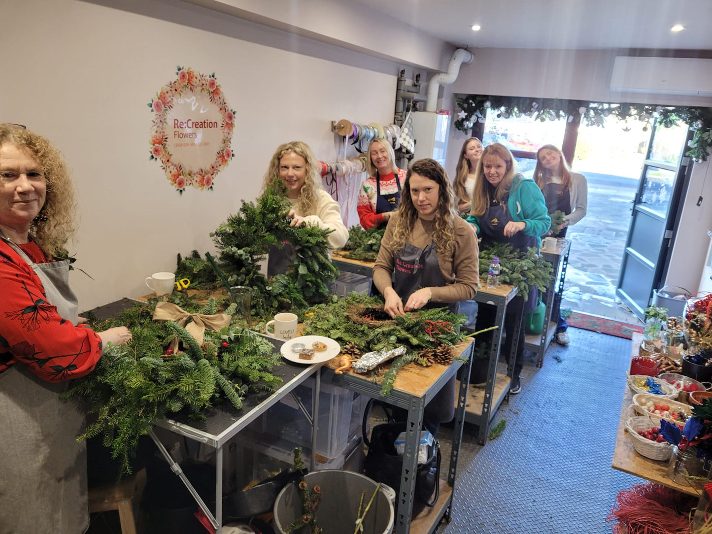 Eight women are creating festive wreaths from greenery at long tables during the Christmas Wreath Class by ReCreation Flowers, smiling and collaborating in a cozy workshop adorned with floral decorations and neatly organized supplies on shelves and tables.
