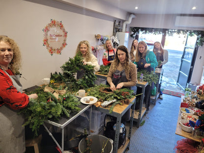 Eight women are creating festive wreaths from greenery at long tables during the Christmas Wreath Class by ReCreation Flowers, smiling and collaborating in a cozy workshop adorned with floral decorations and neatly organized supplies on shelves and tables.