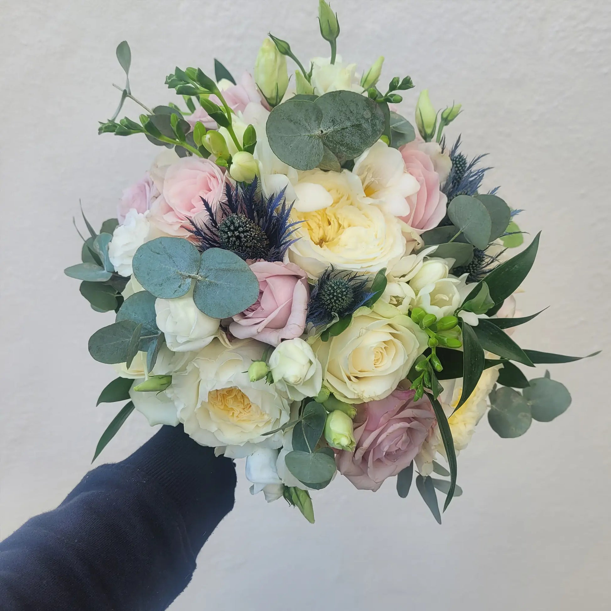 A hand holds a round bouquet of ivory and pale pink roses, white blooms, green eucalyptus, and blue thistle—by ReCreation Flowers—against a plain light background, perfect for the DIY Wedding Flower Package.