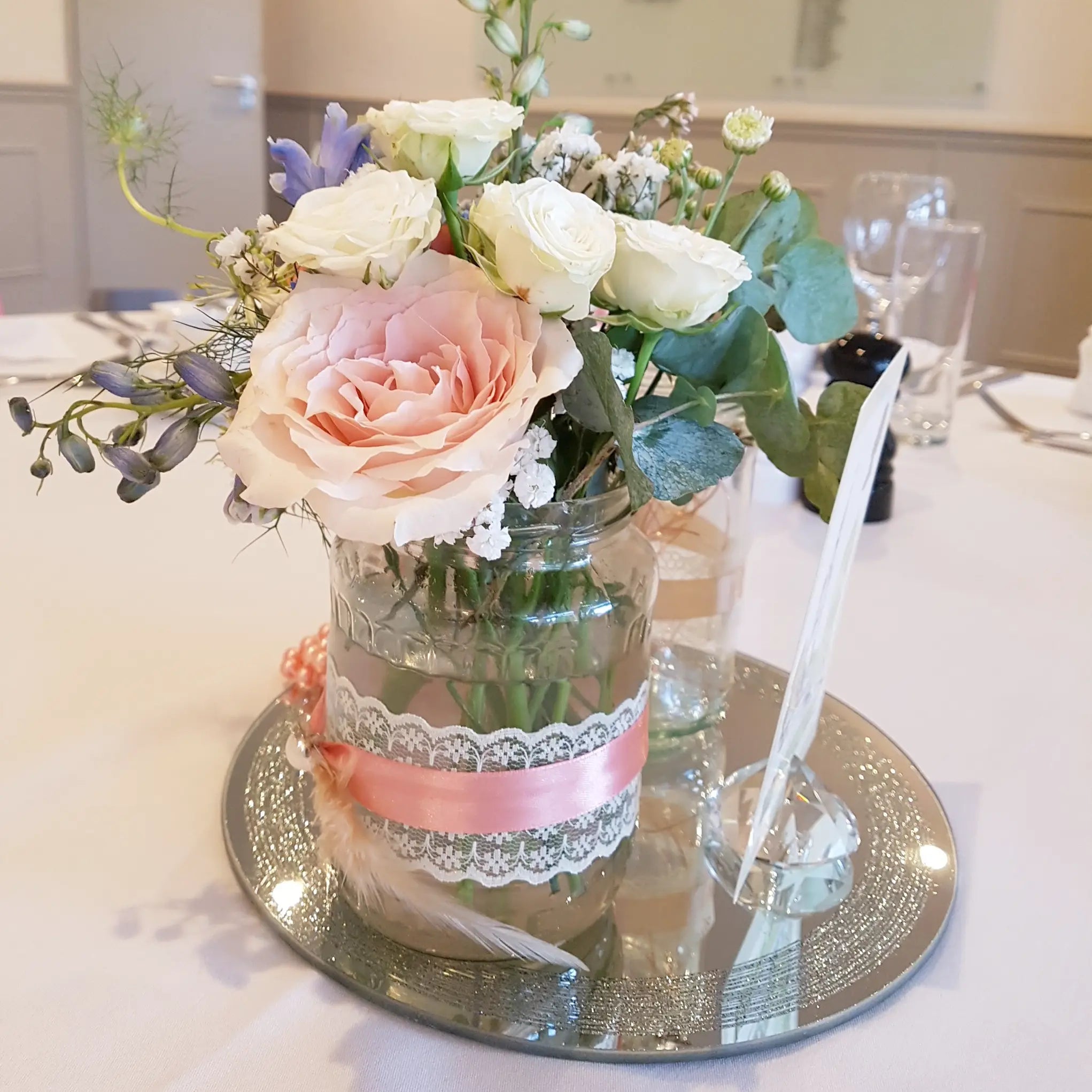 The ReCreation Flowers DIY Wedding Flower Package features a bridal bouquet of roses and greenery in a lace-trimmed jar with a pink ribbon, displayed on a mirrored tray with glasses and a feather.