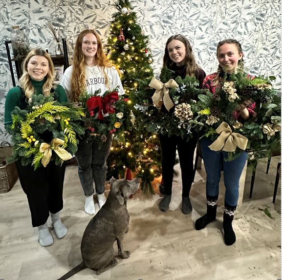 Four women stand indoors by a decorated Christmas tree, each proudly showcasing a festive wreath they made during the ReCreation Flowers Christmas Wreath Class in Leigh on Sea. A gray dog sits at their feet, gazing up at them. Patterned wallpaper and holiday decor in the background perfectly capture the joyful atmosphere of this creative workshop.