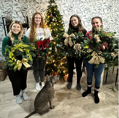 Four women stand indoors by a decorated Christmas tree, each proudly showcasing a festive wreath they made during the ReCreation Flowers Christmas Wreath Class in Leigh on Sea. A gray dog sits at their feet, gazing up at them. Patterned wallpaper and holiday decor in the background perfectly capture the joyful atmosphere of this creative workshop.