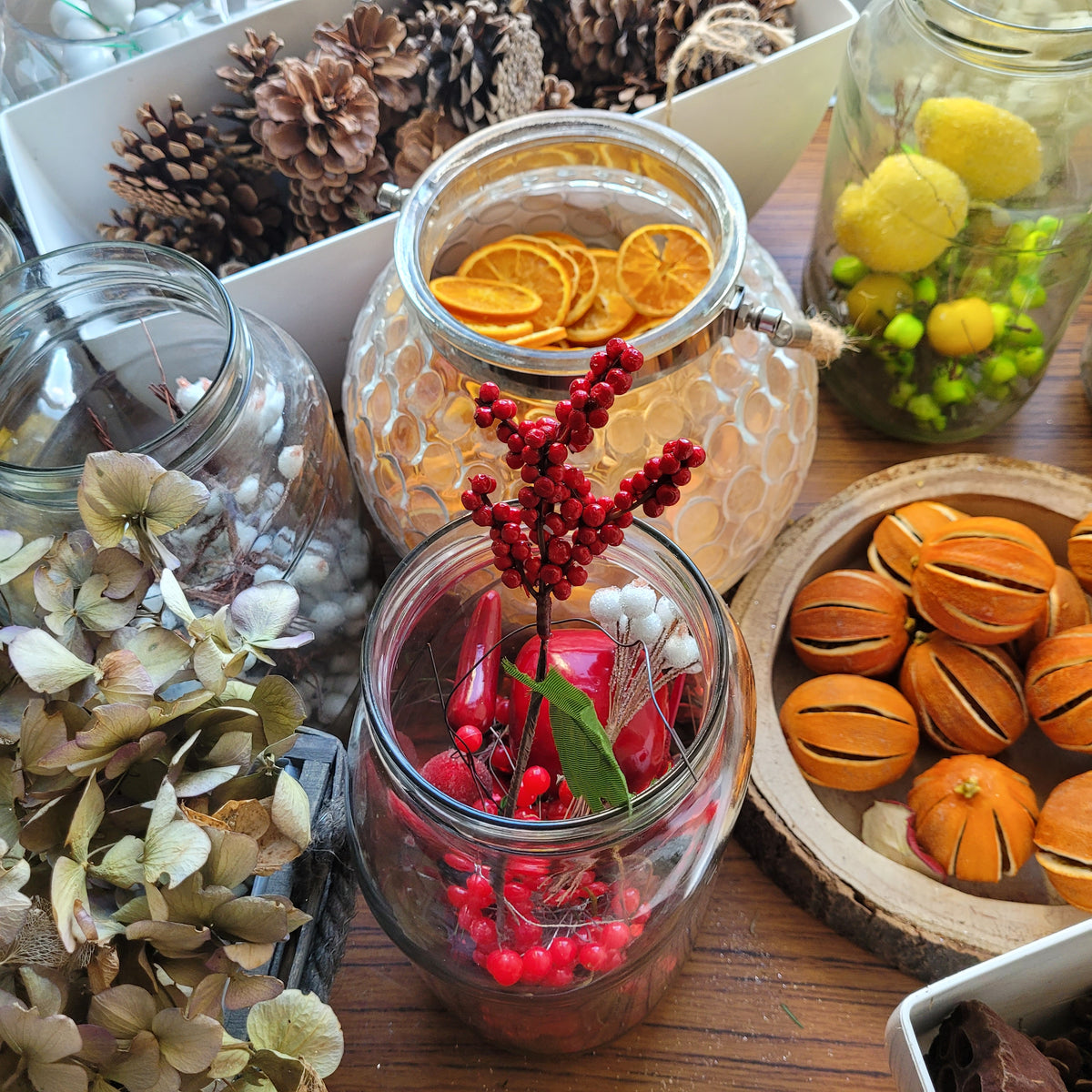 Jars and bowls filled with dried flowers, orange slices, pine cones, and red berries from ReCreation Flowers are beautifully arranged on a wooden table, creating a rustic autumn or holiday decor scene—perfect inspiration for wreath making or candle making at your next Christmas Workshop @ GAS.