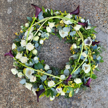 The Funeral Flower Wreath by ReCreation Flowers features white roses, green foliage, and dark purple calla lilies arranged in a circular design with curved stems for a refined funeral tribute.