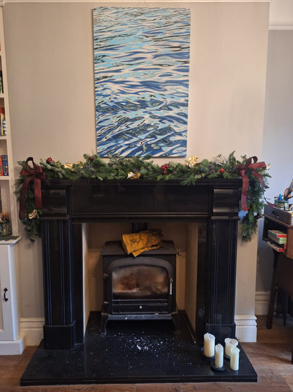 A black fireplace is adorned with a Christmas Garland Class from ReCreation Flowers, featuring fresh foliage, red bows, and gold ornaments. Four candles are arranged on the hearth while a painting of rippling water hangs above the mantle. Bookshelves and a desk frame the cozy scene on each side.