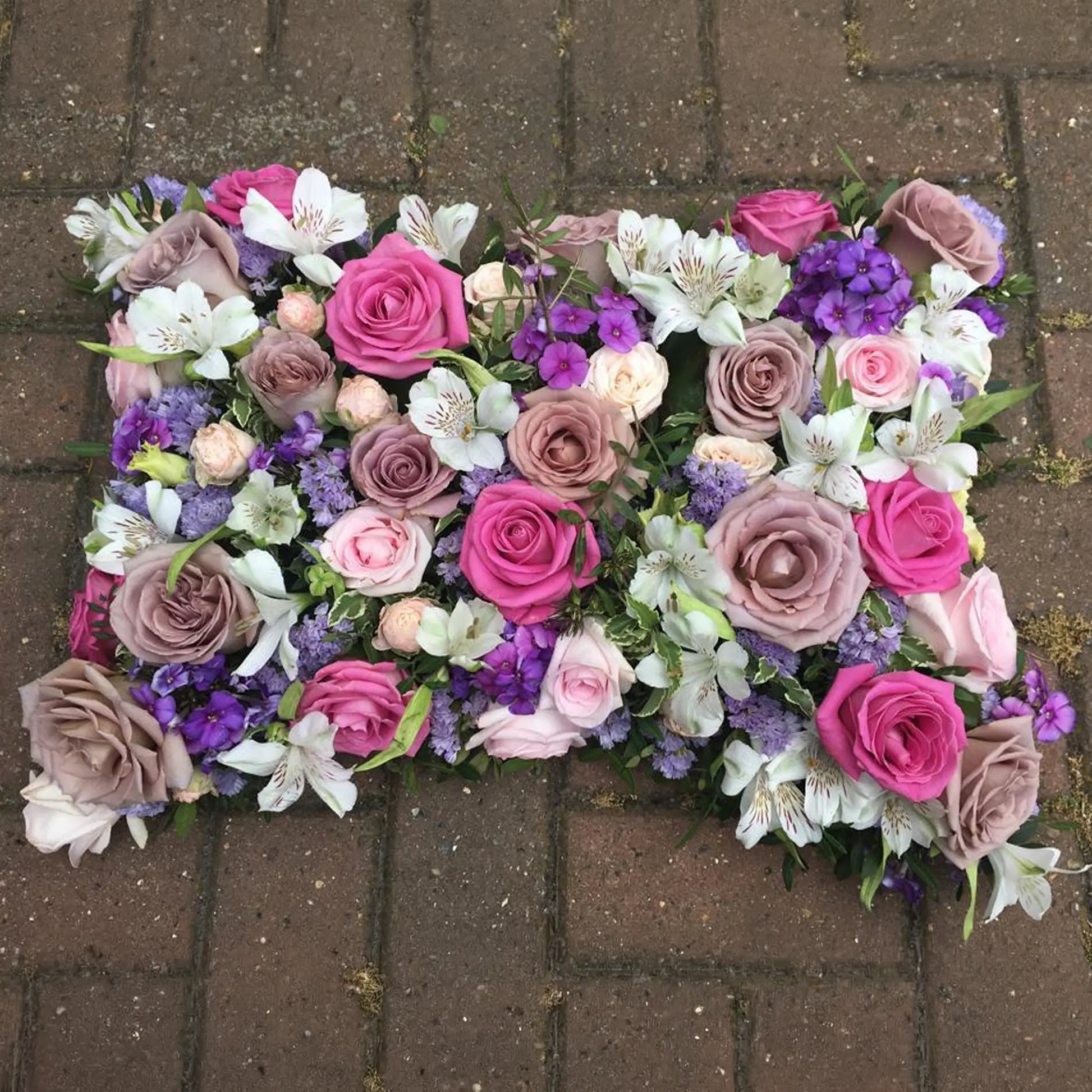 The Cushions & Pillows - Loose Style by ReCreation Flowers features pink, purple, and cream roses, white lilies, and purple seasonal blooms with green foliage arranged on a rectangular tribute pillow.