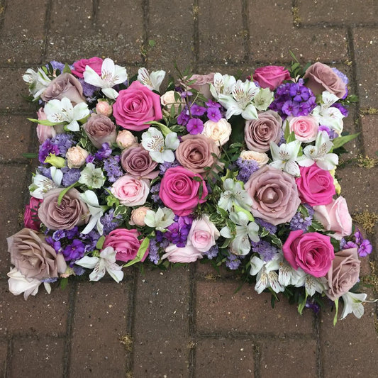 The Cushions & Pillows - Loose Style by ReCreation Flowers features pink, purple, and cream roses, white lilies, and purple seasonal blooms with green foliage arranged on a rectangular tribute pillow.