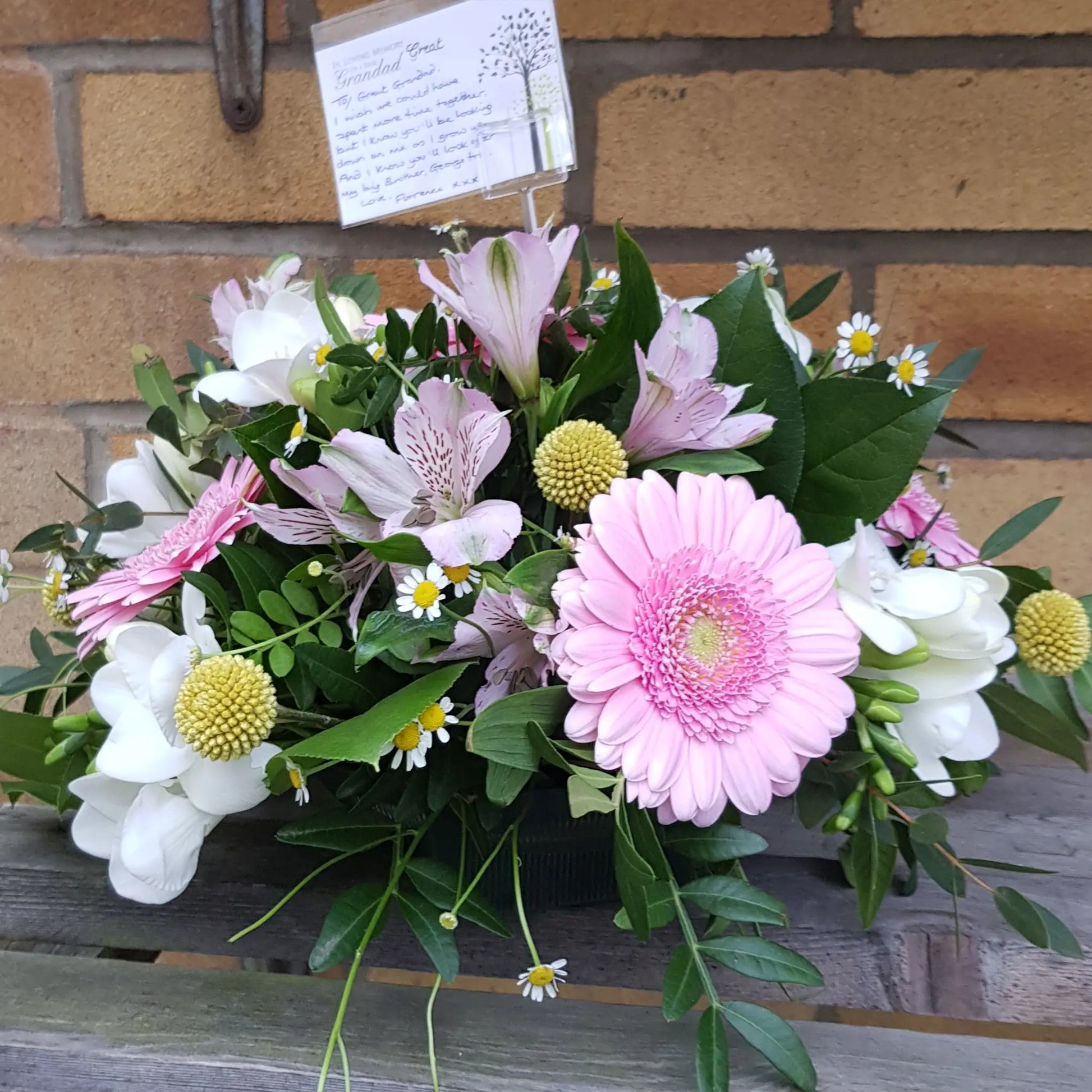 The Funeral Flower Posy by ReCreation Flowers features pink gerbera daisies, white blooms, small daisies, and assorted greenery in a basket, finished with a card at the back.