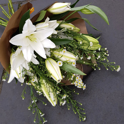The "Scent With Love" bouquet by ReCreation Flowers features white lilies and small daisies with green leaves, wrapped in brown paper and displayed on dark gray paving—a beautiful arrangement from your Leigh-on-Sea florist.