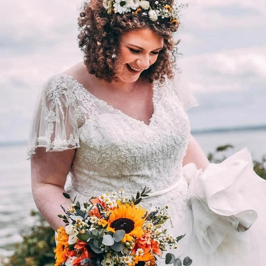 A smiling bride with curly hair and a flower crown stands outdoors near water under a cloudy sky, holding the ReCreation Flowers Seasonal Simplicity Package bridal bouquet of sunflowers and wildflowers.