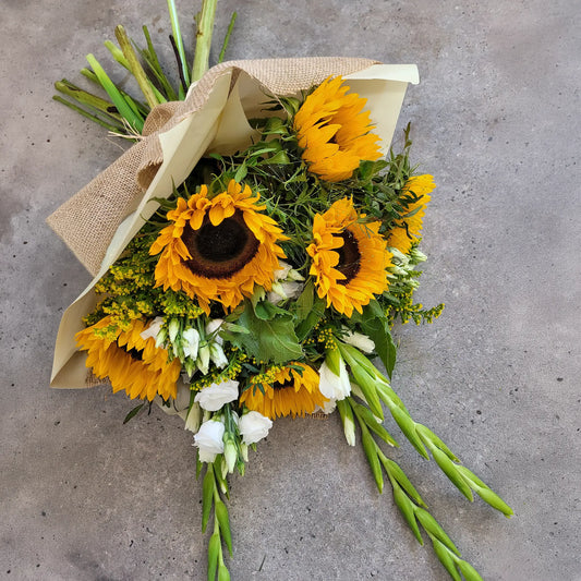 The Funeral Sheaves by ReCreation Flowers feature sunflowers, white blooms, and greenery, artfully wrapped in burlap and cream paper, displayed on a gray textured surface.