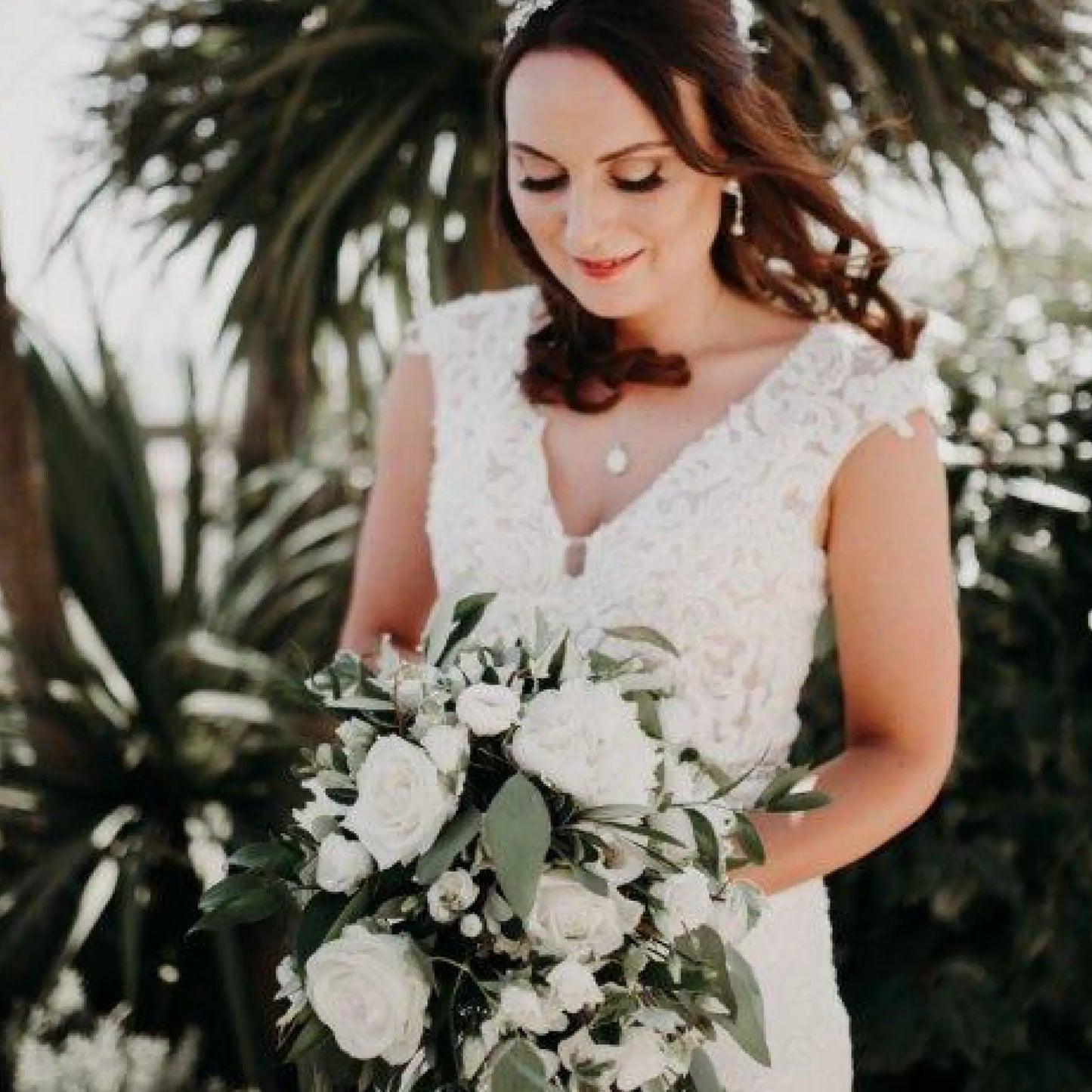 A bride in a white lace gown smiles softly outdoors, holding the Simply Stunning Package by ReCreation Flowers—luxury white roses and greenery bouquet—with palm trees softly blurred in the background.
