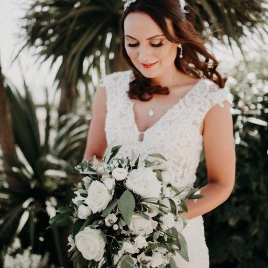 A bride in a white lace gown smiles softly outdoors, holding the Simply Stunning Package by ReCreation Flowers—luxury white roses and greenery bouquet—with palm trees softly blurred in the background.