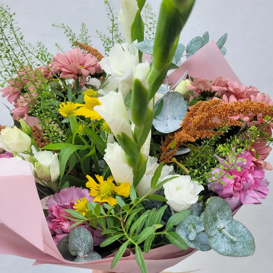 A close-up of a vibrant handmade bouquet by ReCreation Flowers, with white, yellow, pink, and purple blooms in green foliage wrapped in pink—ideal for Flower Subscriptions from ReCreation Flowers.