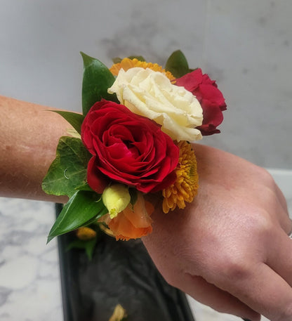 A hand wearing a floral wrist corsage featuring red and white roses, orange and yellow flowers, and green leaves, against a blurred background.