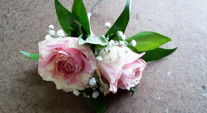 Two pink and white roses with green leaves and small white babys breath flowers are arranged on a brown textured surface.
