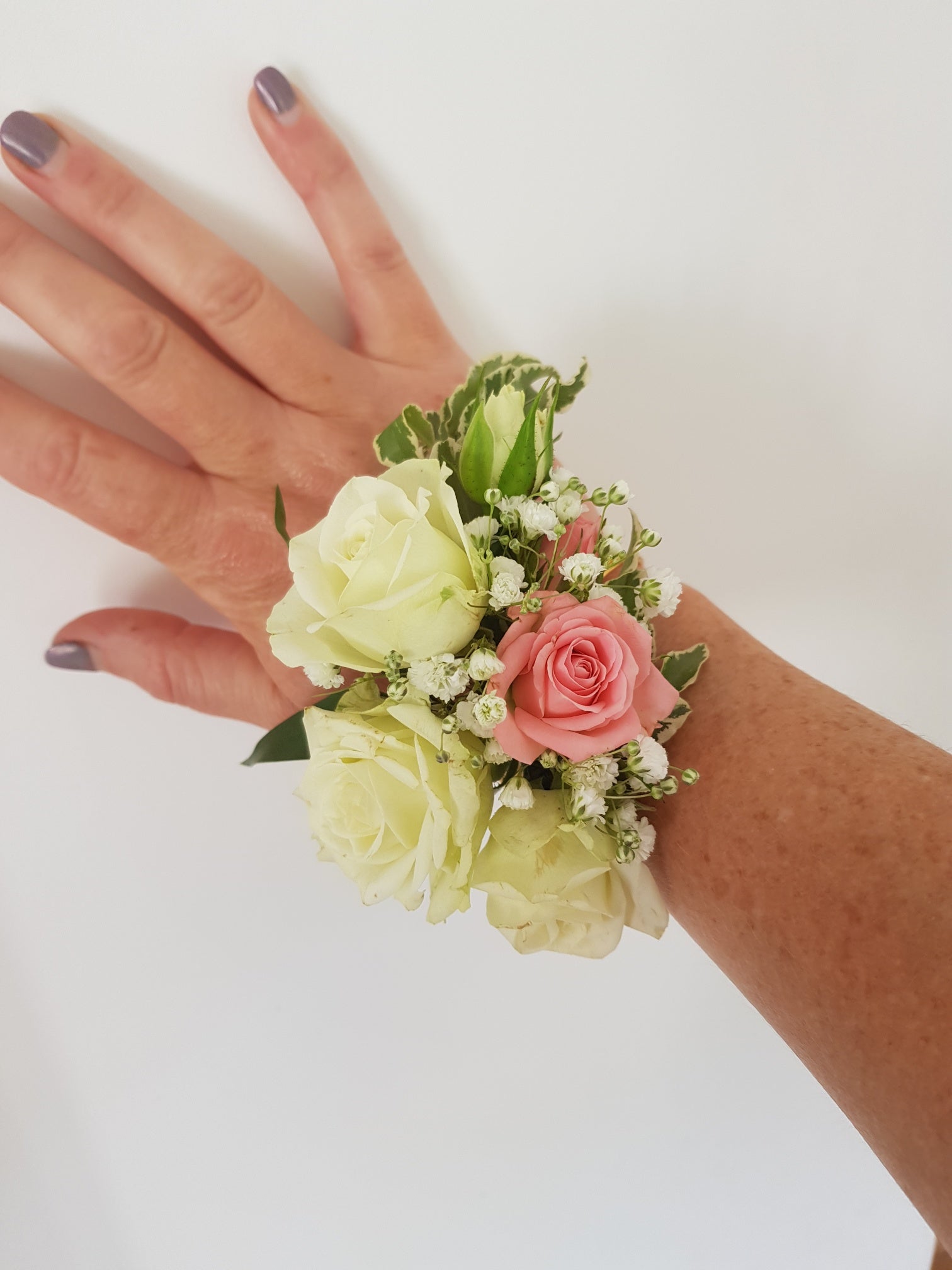 A hand with painted nails wears a floral wrist corsage made of white and pink roses, small white flowers, and green leaves against a plain white background.