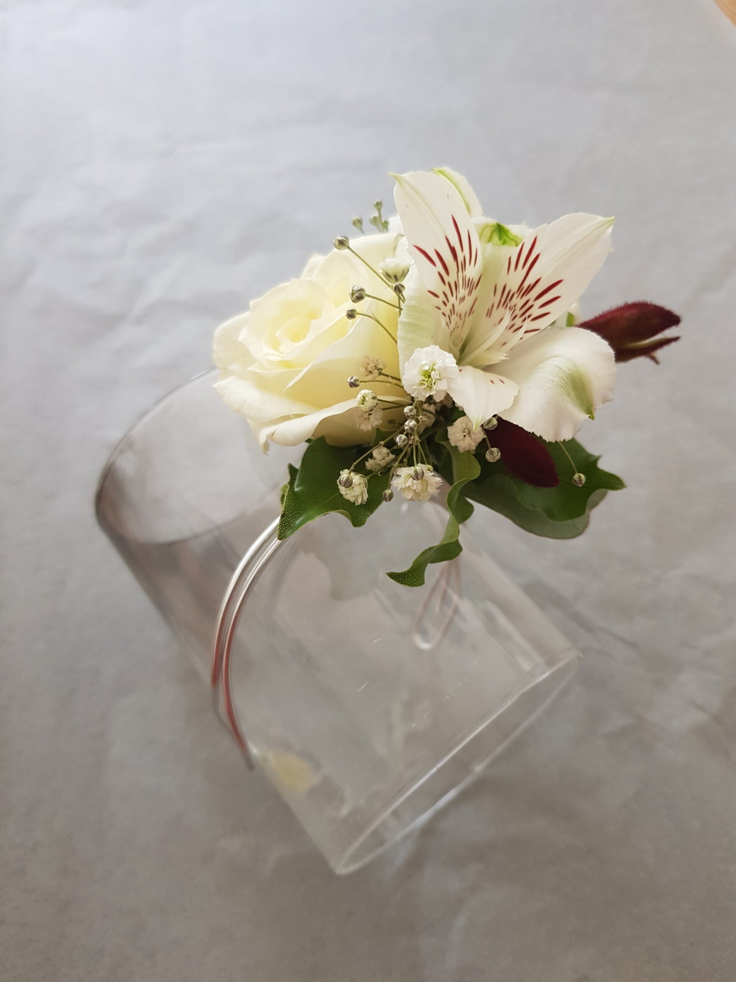 A delicate floral corsage with a white rose, a white lily with red streaks, babys breath, and green leaves, attached to a silver wristband, displayed on a clear glass against a gray background.