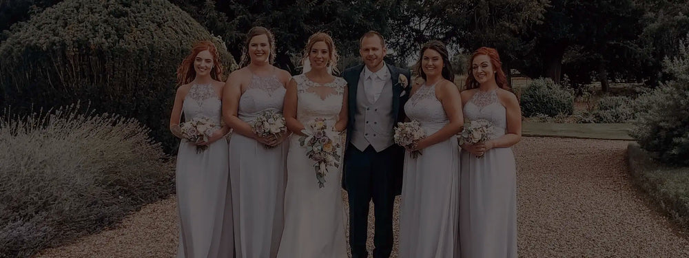 A bride and groom stand outdoors, surrounded by five bridesmaids in matching light gray dresses, all holding bouquets, with trees and greenery in the background.