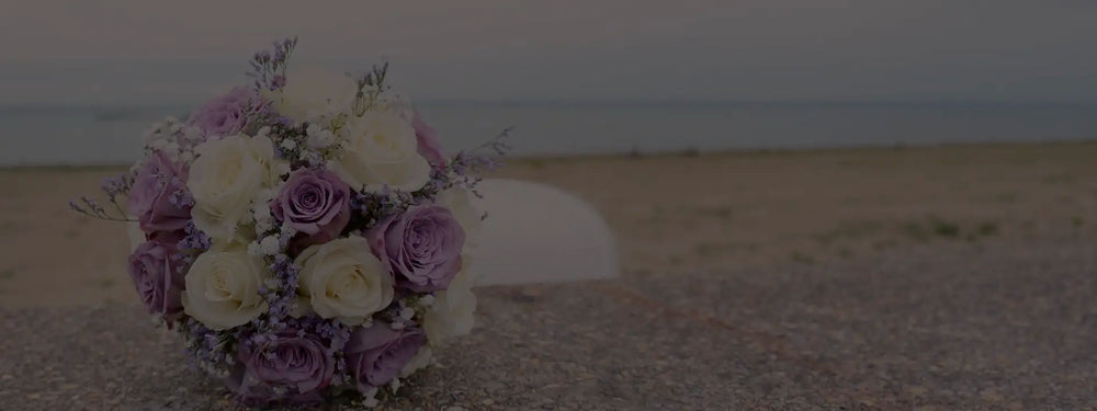 A bouquet of white and light purple roses with small purple flowers rests on a stone surface, with a sandy beach and calm sea in the background.