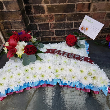 The ReCreation Flowers Cushions & Pillows - White Base is a seasonal tribute pillow with white blooms, red roses, a CHAMPS ribbon, card, and blue-pink trim. Displayed on glass by a brick wall.
