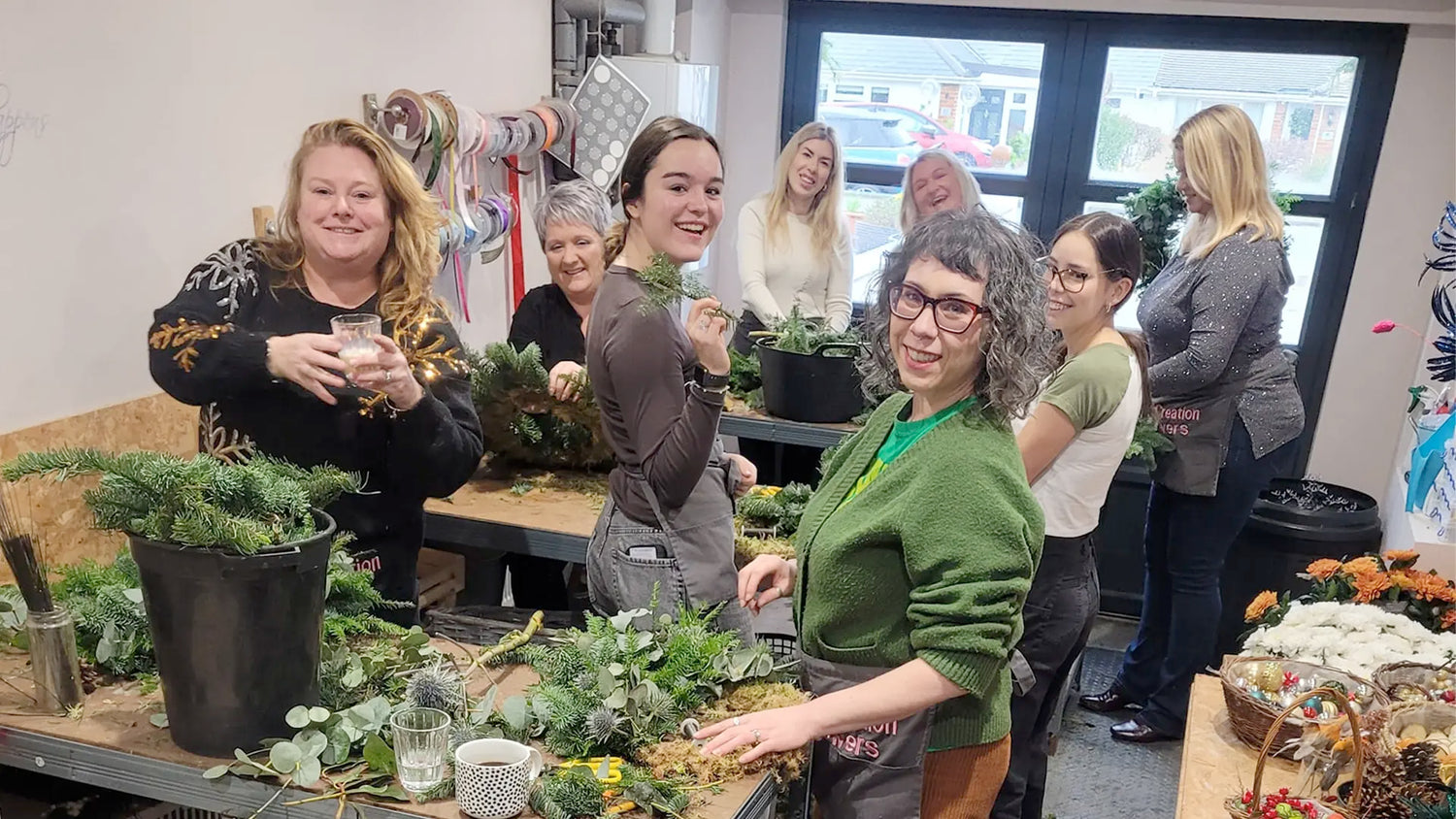 A group of women smiling and arranging greenery and flowers at a workshop, with supplies and plants on tables in a bright, cheerful room.